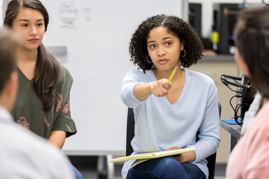 A serious female high school student sits in a circle with a group of classmates and takes a questions as she leads a study group discussion.  She uses her pencil to point to an unrecognizable classmate.